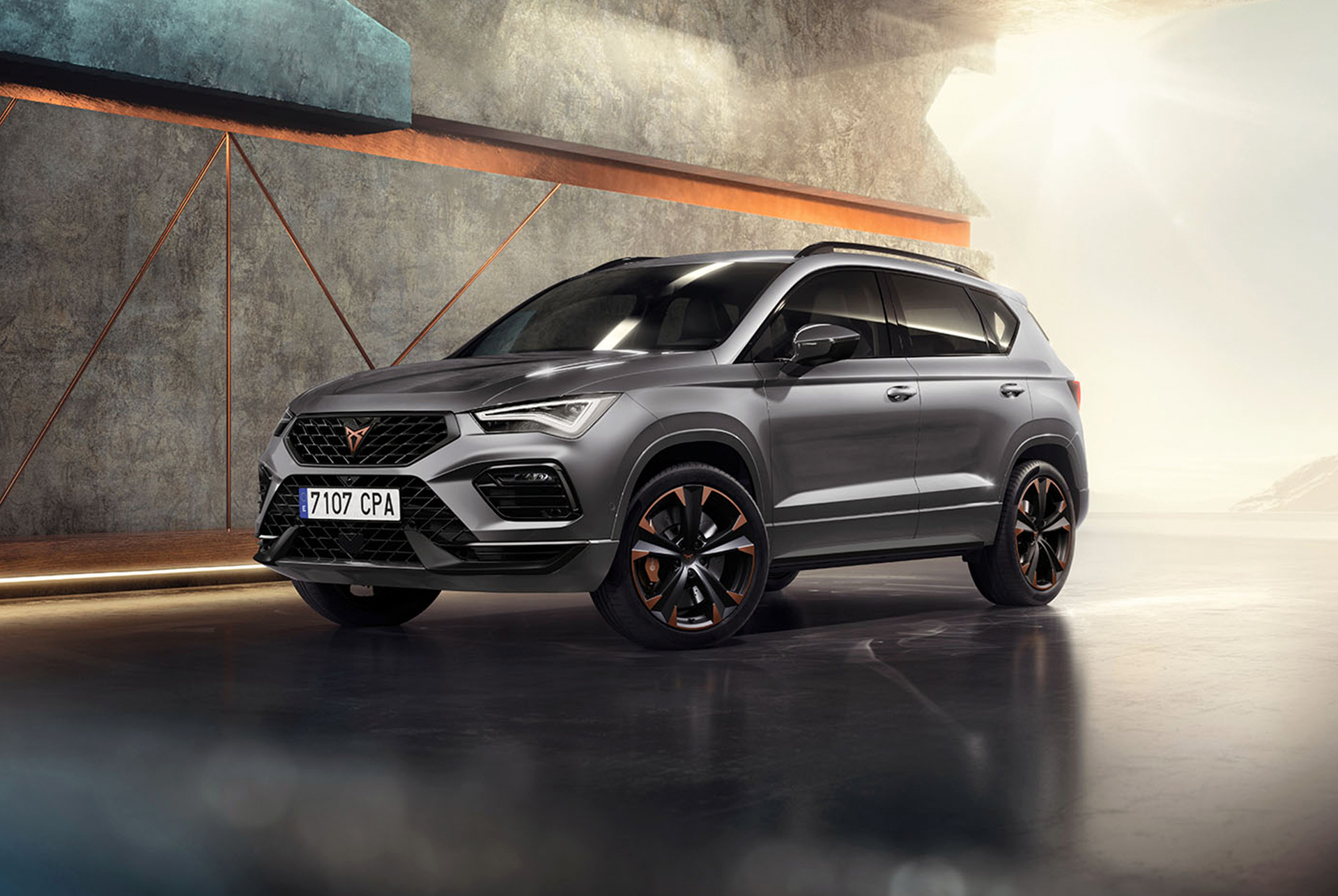 CUPRA Ateca 2024 w kolorze Graphite Grey, sportowy i potężny SUV, zaparkowany w nowoczesnym, industrialnym otoczeniu. Na zdjęciu widać nowoczesną linię nadwozia, reflektory i felgi aluminiowe z elementami w kolorze miedzi. W tle znajduje się ściana o geometrycznych wzorach, które kontrastują z drapieżnym designem auta.