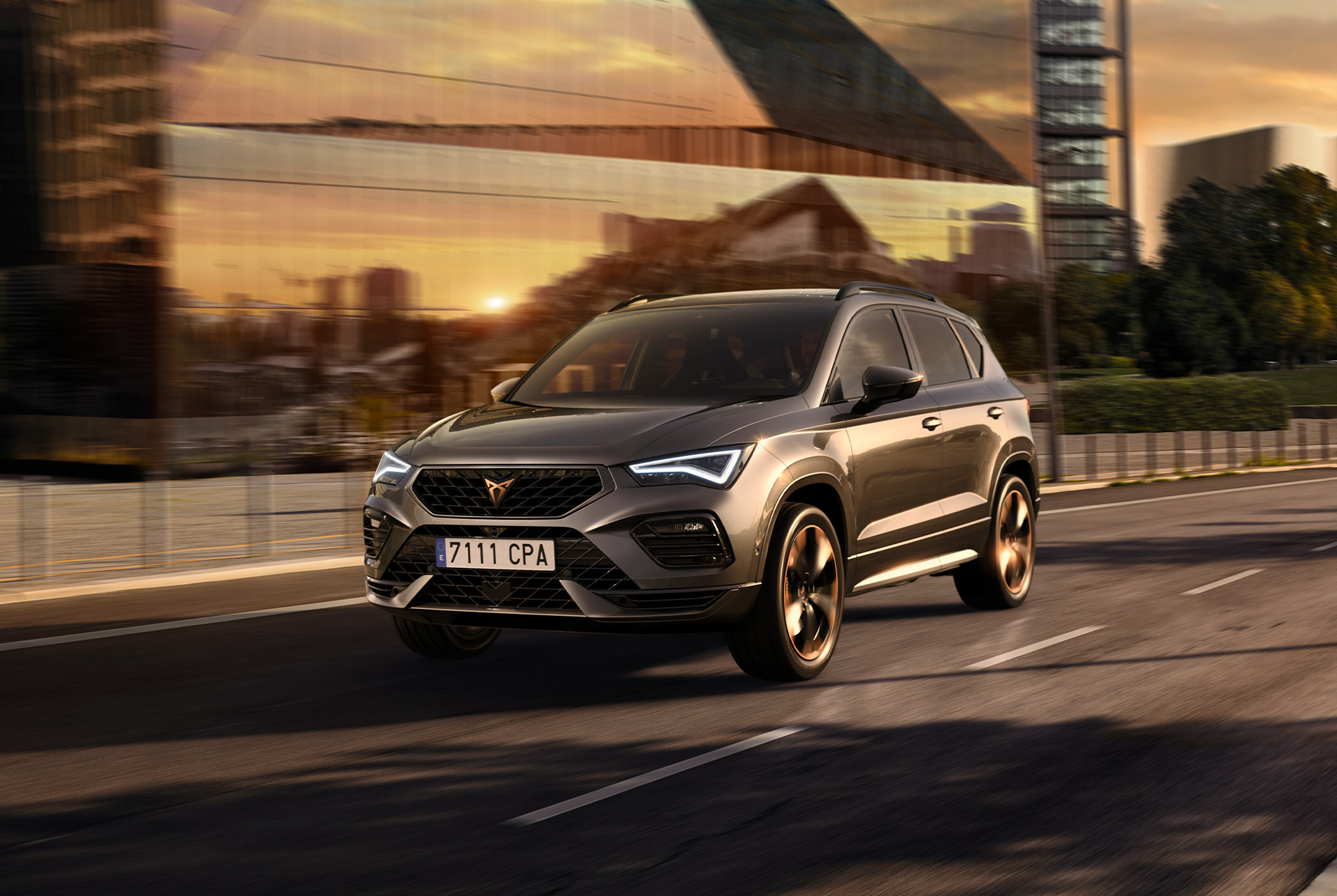 SUV CUPRA Ateca na miejskiej drodze o zachodzie słońca. Dobrze widoczna drapieżna sylwetka nadwozia. Zbliżenie na przedni zderzak, pełne światła LED, maskę oraz logo CUPRA. 