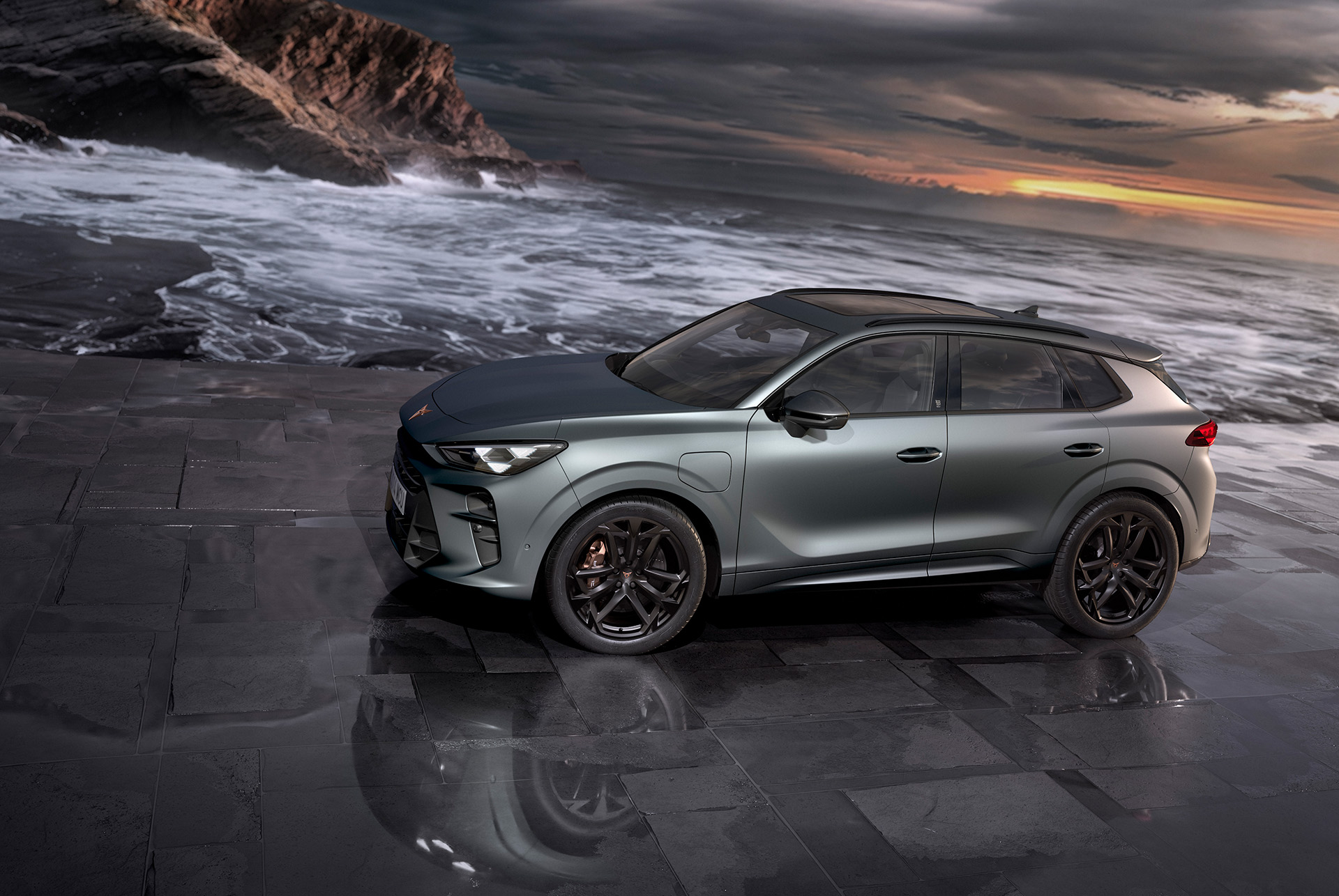 Nowy, hybrydowy SUV CUPRA Terramar 2024 w kolorze Enceladus Grey Matt, wyposażony w 20-calowe felgi aluminiowe America's Cup i hamulce Akebono. Widok na boczną część nadwozia. Auto zaparkowane na mokrym chodniku. W tle widać ocean i zachód słońca.